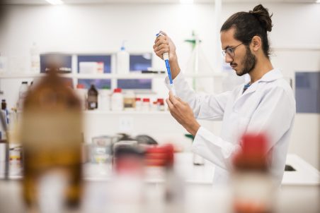 Frascos de laboratório em primeiro plano e um rapaz com jaleco segurando uma pipeta em segundo plano (Foto: Jr. Panela/UFC)