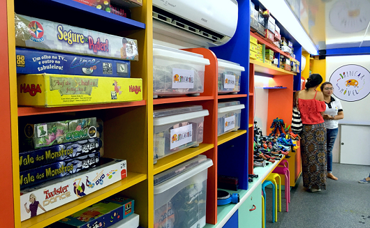 Espaço interior do ônibus com estantes coloridas preenchidas com caixas de brinquedo (Foto: Marília Torres)