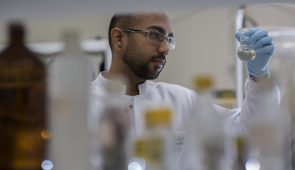 Pesquisador em laboratório segurando um tubo, com medicamentos sem foco em primeiro plano (Foto: Jr. Panela/UFC)