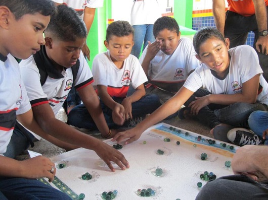 Crianças sentadas no chão em torno de um jogo de tabuleiro com bolas de gude (Foto: Blog Divulgando a Extensão)