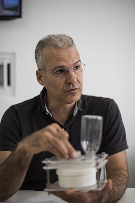 Professor Alberto Nunes sentado à mesa e segurando um equipamento para distribuição de ração (Foto: Jr. Panela/UFC)