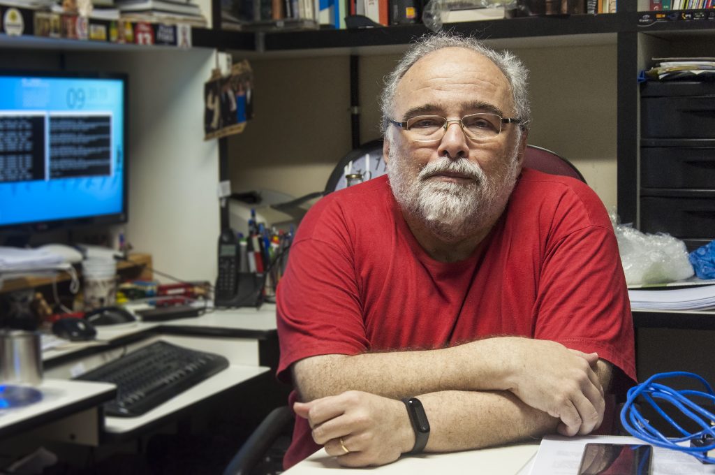 O Prof. Mário Azevedo sendo em posição frontal, em uma sala com livros e um computador (Foto: Viktor Braga/UFC)