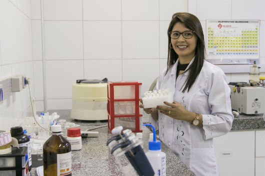 Pesquisadora Suely Alves na bancada do laboratório, segurando frascos com o estudo da liberação do medicamento (Foto: Viktor Braga/UFC)